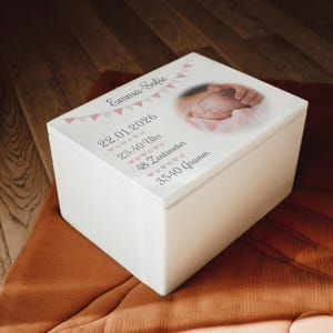 Könnte beinhalten: Eine weiße Andenkenbox mit Deckel, der ein Babyfoto und Text zeigt. Der Deckel ist mit rosa und weißen Wimpeln und Text mit dem Namen des Babys, dem Datum, der Uhrzeit, der Länge (48 cm) und dem Gewicht (3540 Gramm) verziert.