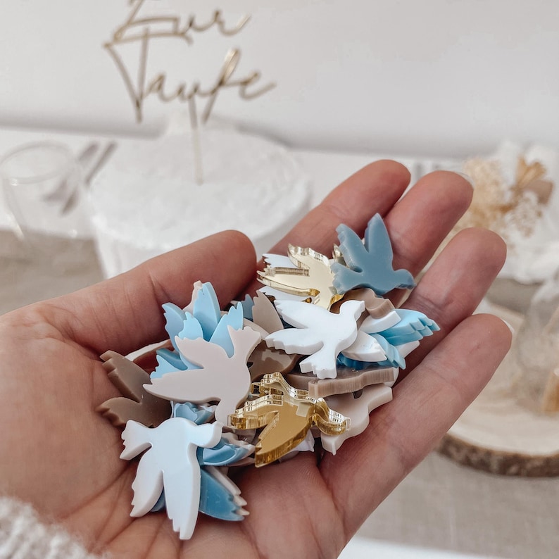 K&ouml;nnte beinhalten: Eine Hand h&auml;lt eine Vielzahl kleiner taubenf&ouml;rmiger Dekorationen. Die Tauben sind aus Acryl und in den Farben Wei&szlig;, Blau, Gold und Braun erh&auml;ltlich.