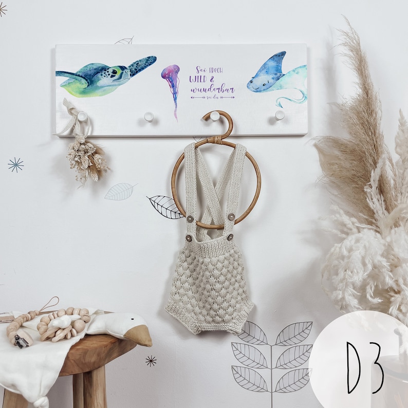May include: White wooden wall-mounted coat rack with three hooks. The rack features a watercolor illustration of a sea turtle, a jellyfish, and a manta ray. The text "See how wild & wunderbar" is printed on the rack. A knitted baby romper hangs on a wooden ring.