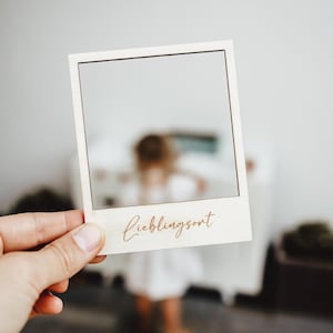 Op de afbeelding: Een wit houten fotolijstje met een uitsparing voor een foto. Het lijstje heeft de tekst "Lieblingsort" erop geschreven in een cursief schrift.