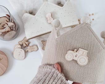 Op de afbeelding: Twee stoffen kronen met pompon accenten. Eén kroon is beige met een houten tractor applicatie, de andere is gebroken wit met een sterrenpatroon. Houten uitsparingen van beren, strikken en cijfers liggen verspreid.
