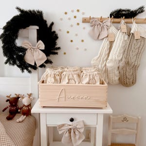 Op de afbeelding: Een houten kist met de naam "Aurora" gegraveerd, gevuld met kleine zakjes met trekkoord. Een zwarte krans met een beige strik hangt boven de kist. Twee gebreide kerstsokken hangen aan de muur. Decoratieve rendieren en een beige strik zijn ook zichtbaar.