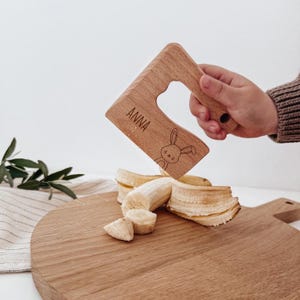 Pode incluir: Um utensílio de cozinha de madeira clara com cabo, gravado com o nome ANNA e um desenho de linha de um coelho. O utensílio está posicionado sobre uma banana descascada numa tábua de cortar redonda de madeira com grão natural.