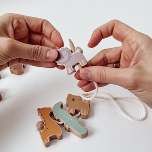 Könnte beinhalten: Holzspielzeug-Set mit Tieren: Löwe, Giraffe, Krokodil und Bär. Der Löwe ist helllila, die Giraffe orange und das Krokodil hellblau. Die Tiere sind durch eine weiße Schnur verbunden. Das Spielzeug wird von zwei Händen gehalten.