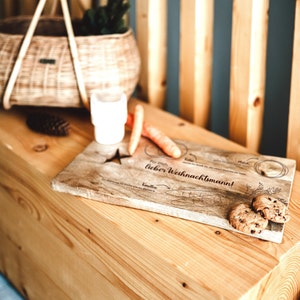May include: A wooden cutting board with the text "F&uuml;r Dich, lieber Weihnachtsmann!" and a drawing of a Christmas tree, a star, and a carrot. There are also two cookies on the board.