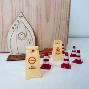 Könnte beinhalten: Eine Miniatur-Holztür mit Herzausschnitt und Sternengravuren, begleitet von kleinen Kunststoff-Bauzubehörteilen, darunter zwei gelbe Warnschilder mit Text und vier rot-weiße Verkehrskegel.