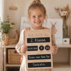 Könnte beinhalten: Ein rechteckiges Meilensteintafel aus Holz mit schwarzen Tafelflächen, beschriftet mit 'MEIN ERSTER TAG', '2. Klasse', '03.09.2026', '7', '120', 'Tanzen, mein Hase Fips' und 'Tierärztin', verziert mit einer eingravierten Wimpelkette.