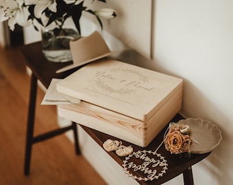 Puede incluir: Una caja de madera con tapa, grabada con las palabras "Natalie & Pascal" en una fuente cursiva. La caja está decorada con una corona de hojas y flores. La caja está sentada sobre una mesa de madera con un sombrero, un ramo de flores y un plato pequeño con un anillo.