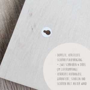 May include: A close-up of a light wood surface with a white circle containing a hole. Text in German reads: "Doppelte, versteckte Sicherheitsaufh&auml;ngung + Zwei Schrauben & D&uuml;bel (im Lieferumfang) - Verdrehte Aufh&auml;ng&ouml;se, garantiert, stabilen und sicheren Halt an der Wand"