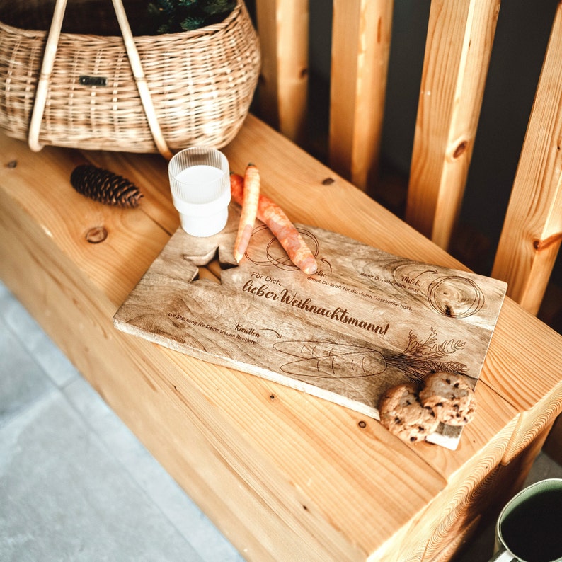 May include: A wooden serving board with the text "For you, dear Santa Claus!" engraved on it. The board is decorated with drawings of carrots, milk, and cookies. There are also two cookies on the board.