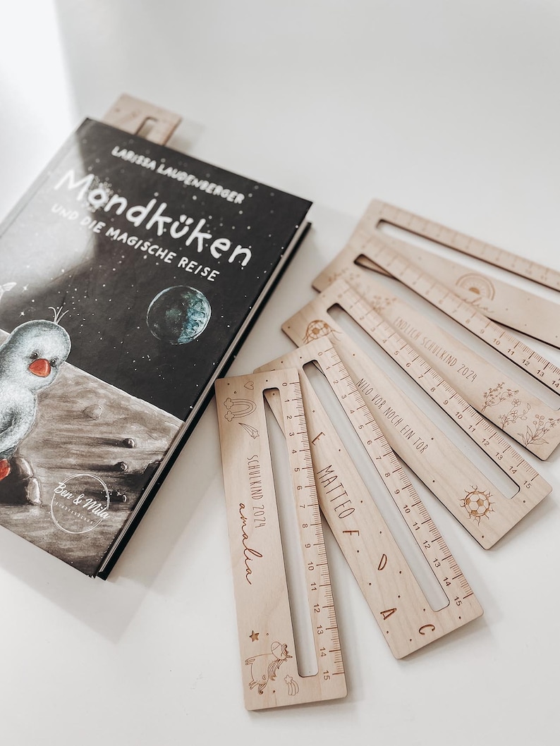 K&ouml;nnte beinhalten: Ein Set aus f&uuml;nf Holzlinealen mit personalisierten Namen und Skalierungen. Jedes Lineal hat ein einzigartiges Design, darunter ein Einhorn, ein Fu&szlig;ball, ein Regenbogen und eine Blume. Die Lineale sind aus Naturholz gefertigt und haben eine hellbraune Farbe.