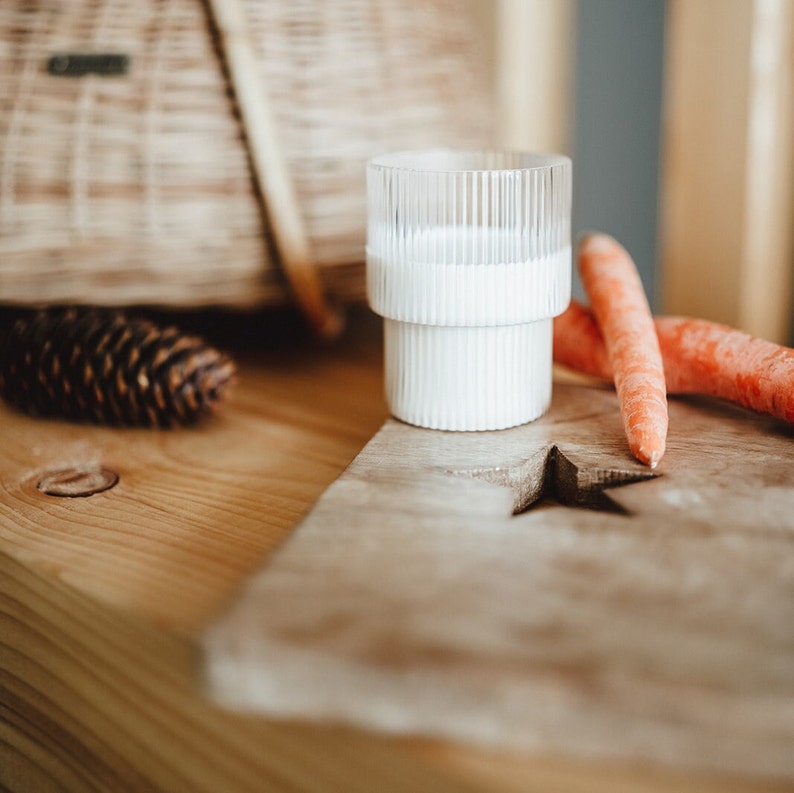 May include: A white glass with a ribbed design filled with a white liquid sits on a wooden cutting board with two orange carrots. A star shape is carved into the board.