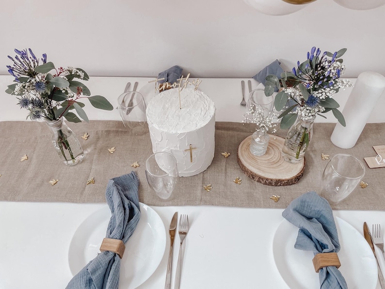 K&ouml;nnte beinhalten: Ein wei&szlig;er Kuchen mit einem goldenen Kreuz-Topper und dem Namen "Timothy" obenauf, auf einem Tisch mit blauen Servietten, goldenen Konfetti und zwei Vasen mit blauen Blumen.