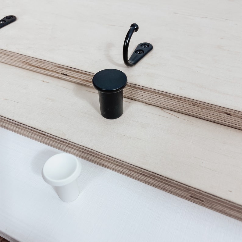 May include: Two cabinet knobs, one black and one white, are shown on a light wood surface. A black metal hook is also visible.