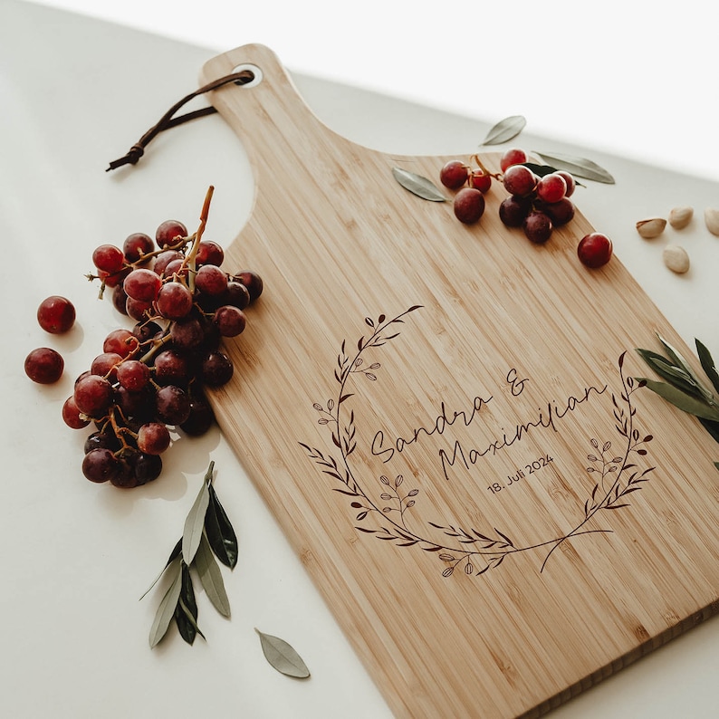 Puede incluir: Una tabla de cortar de bamb&uacute; personalizada con un dise&ntilde;o de corona floral y el texto "Sandra & Maximilian 18. Juli 2024".