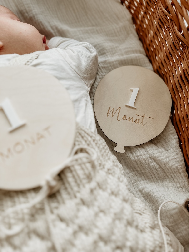 Puede incluir: Un letrero de madera para beb&eacute; en forma de globo con el n&uacute;mero "1" y la palabra "Monat" escrita en &eacute;l. El letrero es beige con un n&uacute;mero y un texto blancos.