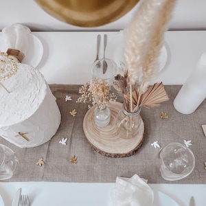 K&ouml;nnte beinhalten: Ein wei&szlig;er Kuchen mit goldenen Akzenten steht auf einem Tisch mit einer braunen Leinentischdecke. Ein Strau&szlig; getrockneter Blumen und Pampasgras steht in einer Glasvase auf einer Holzscheibe. Wei&szlig;e Tauben sind auf dem Tisch verstreut.