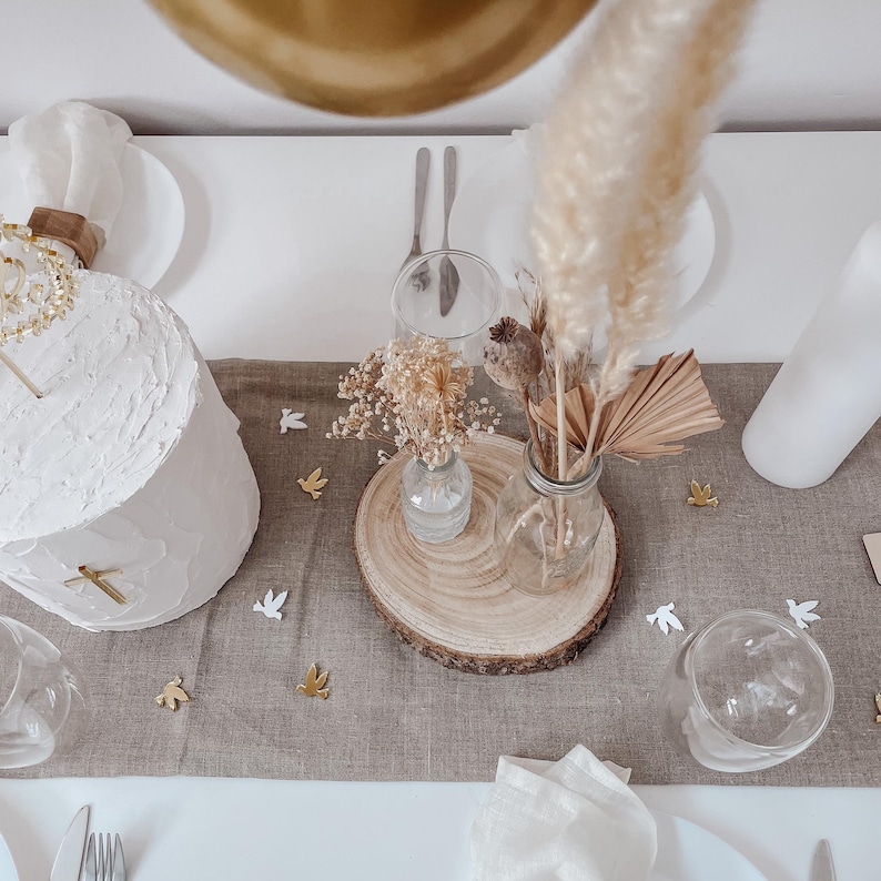 K&ouml;nnte beinhalten: Ein wei&szlig;er Kuchen mit goldenen Akzenten steht auf einem Tisch mit einer braunen Leinentischdecke. Ein Strau&szlig; getrockneter Blumen und Pampasgras steht in einer Glasvase auf einer Holzscheibe. Wei&szlig;e Tauben sind auf dem Tisch verstreut.