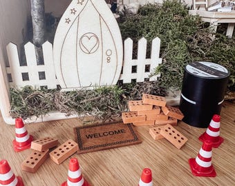 Può includere: Una scena di giardino delle fate in miniatura con una porta di legno, una staccionata bianca e uno zerbino. Include piccoli mattoni arancioni, coni stradali rossi e bianchi e una bobina di filo nero. La porta ha un design a cuore e stella.