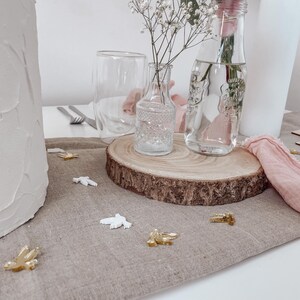 K&ouml;nnte beinhalten: Eine Holzscheibe mit einer durchsichtigen Glasvase gef&uuml;llt mit wei&szlig;en Babyschnapsblumen und einer durchsichtigen Glasflasche gef&uuml;llt mit Wasser. Die Holzscheibe liegt auf einer beigefarbenen Leinentischdecke mit wei&szlig;en und goldenen Dekorationsv&ouml;geln, die um sie herum verstreut sind.