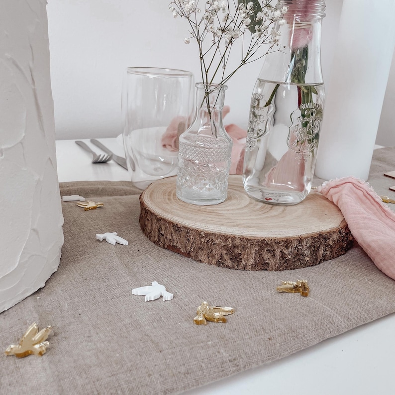 K&ouml;nnte beinhalten: Eine Holzscheibe mit einer durchsichtigen Glasvase gef&uuml;llt mit wei&szlig;en Babyschnapsblumen und einer durchsichtigen Glasflasche gef&uuml;llt mit Wasser. Die Holzscheibe liegt auf einer beigefarbenen Leinentischdecke mit wei&szlig;en und goldenen Dekorationsv&ouml;geln, die um sie herum verstreut sind.