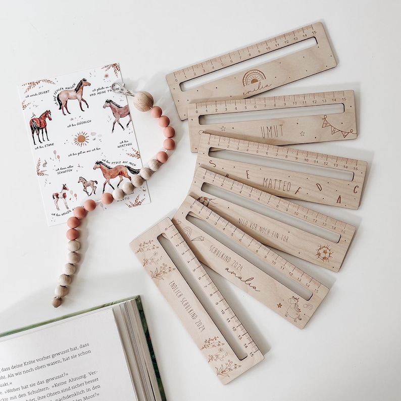 K&ouml;nnte beinhalten: Sechs Holzlineale mit verschiedenen Designs und Namen. Die Lineale sind alle 15 Zoll lang und haben einen Ausschnitt in der Mitte. Die Designs beinhalten einen Regenbogen, einen Stern, einen Fu&szlig;ball, ein Einhorn und ein Blumenmuster. Die Namen auf den Linealen sind "Emilia", "Umut", "Matteo", "Nilo", "Amalia" und "Endlich Schulkind 2023".