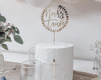 Op de afbeelding: Gouden taarttopper met een krans van bladeren en de tekst "Noah's Taufe" op een witte taart met een gouden kruis.