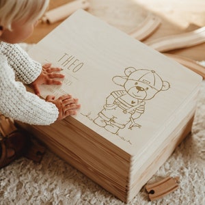 Peut inclure: Un coffre à jouets en bois avec une finition en bois clair. Le couvercle porte une inscription dorée qui dit "Theo 2023" et représente un ours en dessin animé portant une casquette de baseball et une salopette.