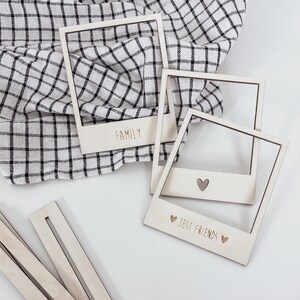 Op de afbeelding: Drie houten fotolijsten op een witte achtergrond. De lijsten zijn in de vorm van een Polaroid-camera. De bovenste lijst zegt "FAMILY" in een gouden lettertype. De middelste lijst is leeg. De onderste lijst zegt "BEST FRIENDS" in een gouden lettertype met twee harten aan weerszijden van de tekst.