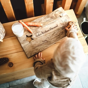 May include: A wooden cutting board with a star shape cut out. The board is engraved with the text "For you, dear Santa Claus!" and "So that you have enough for all the presents". There is a carrot, a cookie, and a glass of milk on the board. A child is reaching for the cookie.