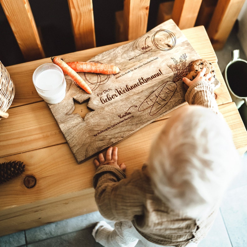 May include: A wooden cutting board with a star shape cut out. The board is engraved with the text "For you, dear Santa Claus!" and "So that you have enough for all the presents". There is a carrot, a cookie, and a glass of milk on the board. A child is reaching for the cookie.