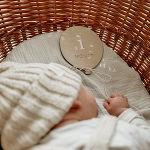 Puede incluir: Un beb&eacute; con un gorro de punto blanco duerme en una cesta de mimbre. Un c&iacute;rculo de madera con el n&uacute;mero "1" y la palabra "MONAT" se coloca sobre la tela blanca en la cesta.