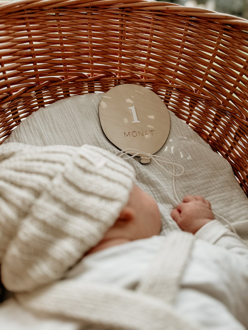 Puede incluir: Un beb&eacute; con un gorro de punto blanco duerme en una cesta de mimbre. Un c&iacute;rculo de madera con el n&uacute;mero "1" y la palabra "MONAT" se coloca sobre la tela blanca en la cesta.