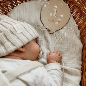 Puede incluir: Un beb&eacute; durmiendo en una cesta de mimbre con un letrero de madera que dice "1 Monat". El letrero tiene forma de globo y tiene una cuerda atada.