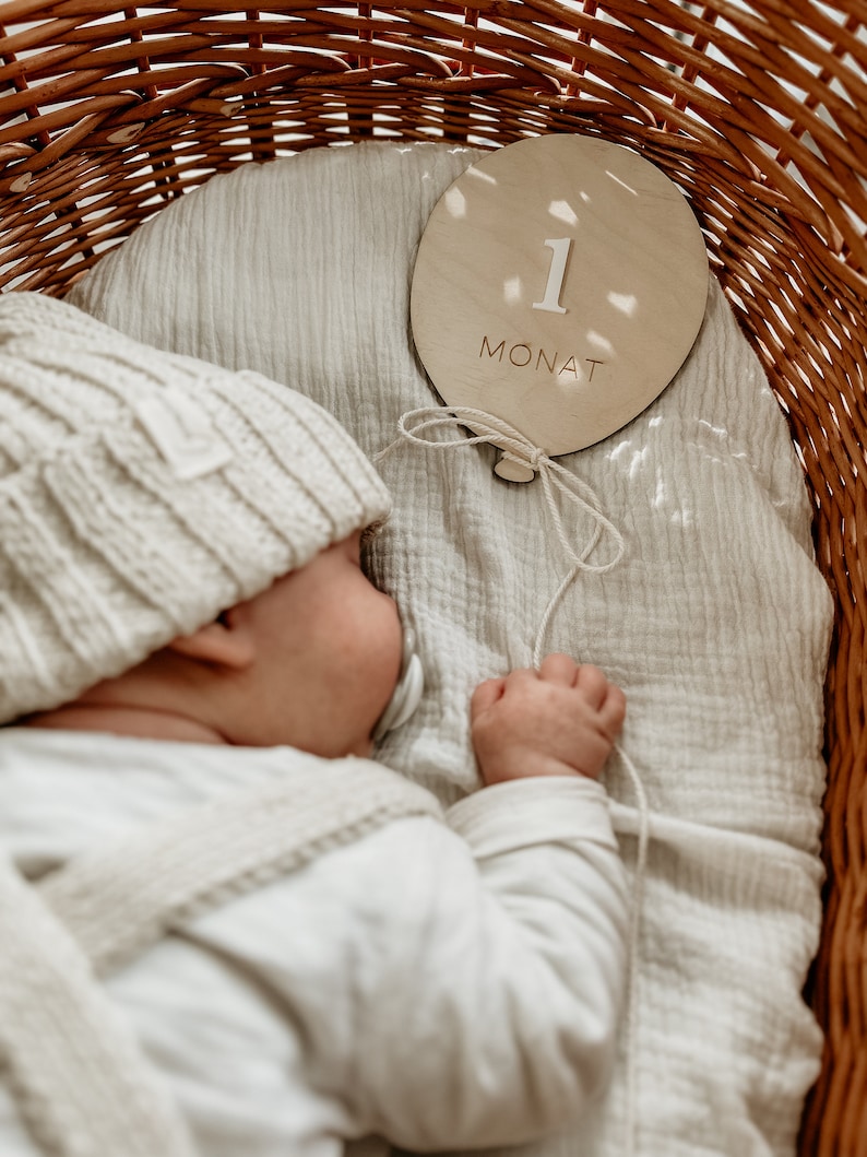 Puede incluir: Un beb&eacute; durmiendo en una cesta de mimbre con un letrero de madera que dice "1 Monat". El letrero tiene forma de globo y tiene una cuerda atada.
