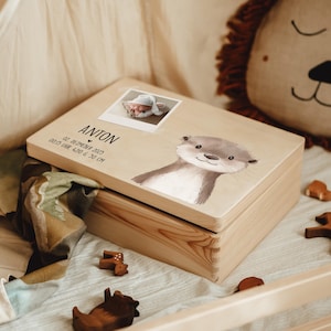Op de afbeelding: Een lichtbruine houten herinneringsdoos. De deksel toont een babyfoto, de naam "ANTON" en geboortegegevens. Een illustratie van een otter staat rechts. De doos is omgeven door een deken en houten speelgoed. De baby was 50 cm lang.