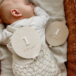 Puede incluir: Un beb&eacute; con un atuendo de punto de color crema con un letrero de madera que dice "1 Monat" sujeto a &eacute;l. El beb&eacute; est&aacute; durmiendo en una cesta de mimbre.