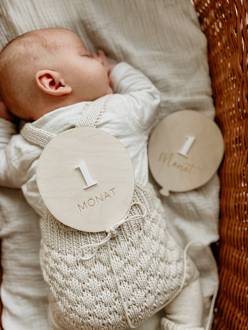Puede incluir: Un beb&eacute; con un atuendo de punto de color crema con un letrero de madera que dice "1 Monat" sujeto a &eacute;l. El beb&eacute; est&aacute; durmiendo en una cesta de mimbre.