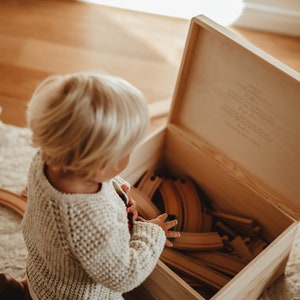 Ein kleiner Junge vor unserer Holzkiste mit Spielzeug drin. Im Innendeckel ein wunderschöner Spruch. Unsere Hände werden dich halten.  In der Kiste können auch hochwertige Erinnerungen aufbewahrt werden.