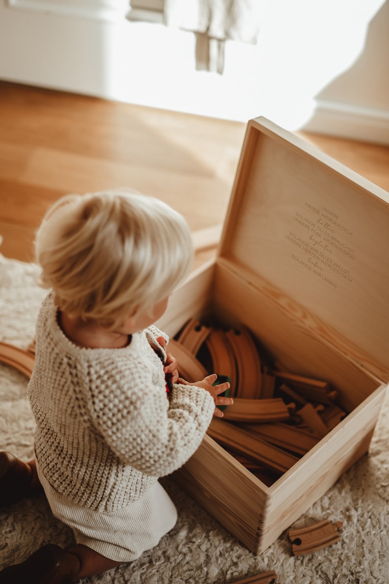 Ein kleiner Junge vor unserer Holzkiste mit Spielzeug drin. Im Innendeckel ein wunderschöner Spruch. Unsere Hände werden dich halten.  In der Kiste können auch hochwertige Erinnerungen aufbewahrt werden.