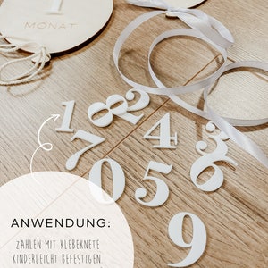 Puede incluir: N&uacute;meros blancos del 1 al 9 con respaldo adhesivo para usar como un rastreador de hitos para beb&eacute;s. Los n&uacute;meros est&aacute;n sobre una superficie de madera con dos c&iacute;rculos de madera blancos con los n&uacute;meros 1 y las palabras "1 Monat" y "1 Monate" escritas en ellos. Una cinta blanca est&aacute; envuelta alrededor de los c&iacute;rculos.