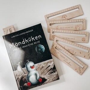 K&ouml;nnte beinhalten: Ein schwarz-wei&szlig; illustriertes Kinderbuch mit dem Titel "Mondk&uuml;ken und die magische Reise" von Larissa Laudenberger. Der Buchumschlag zeigt einen Comic-Pinguin auf dem Mond. Das Buch wird von f&uuml;nf Holzlinealen mit personalisierten Namen und Daten begleitet.