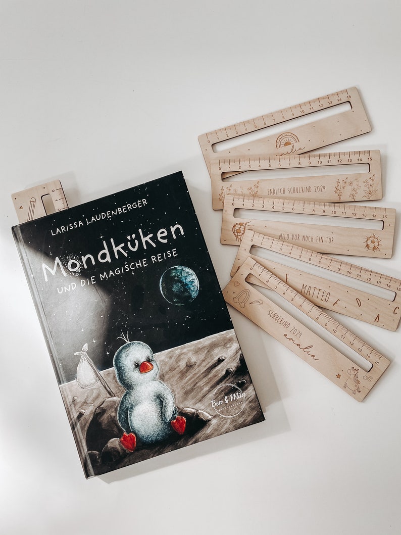 K&ouml;nnte beinhalten: Ein schwarz-wei&szlig; illustriertes Kinderbuch mit dem Titel "Mondk&uuml;ken und die magische Reise" von Larissa Laudenberger. Der Buchumschlag zeigt einen Comic-Pinguin auf dem Mond. Das Buch wird von f&uuml;nf Holzlinealen mit personalisierten Namen und Daten begleitet.