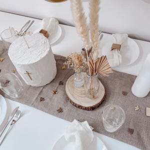 K&ouml;nnte beinhalten: Ein wei&szlig;er Kuchen mit einem goldenen Kreuz und dem Wort "Mama" obenauf, dekoriert mit getrockneten Blumen und Pampasgras in Glasvasen auf einem Holzscheiben. Der Kuchen steht auf einer braunen Leinen-Tischdecke mit goldenen Konfetti.