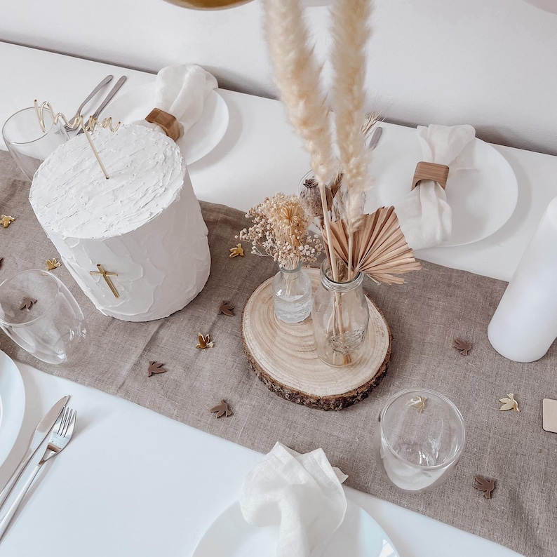 K&ouml;nnte beinhalten: Ein wei&szlig;er Kuchen mit einem goldenen Kreuz und dem Wort "Mama" obenauf, dekoriert mit getrockneten Blumen und Pampasgras in Glasvasen auf einem Holzscheiben. Der Kuchen steht auf einer braunen Leinen-Tischdecke mit goldenen Konfetti.