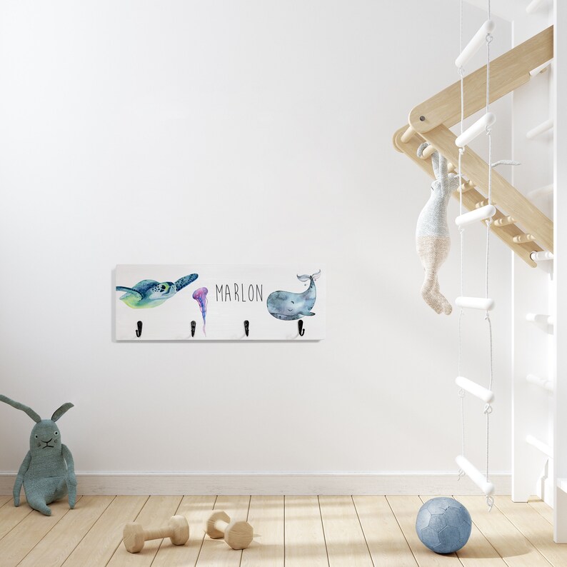 May include: White wooden coat rack with four hooks. The rack features a watercolor design of a sea turtle, jellyfish, and whale. The name "MARLON" is printed on the rack.