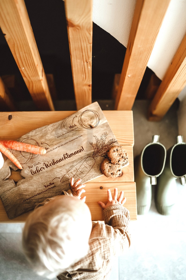 May include: A wooden cutting board with a hand-drawn illustration of a carrot and the text "For you, dear Santa Claus!" in German. The board is decorated with a green sprig of wheat and a small circle with the word "Milk" written inside. The board is sitting on a wooden table with two green rubber boots in the background.