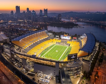 May include: An aerial view of a large, illuminated football stadium with a green field and yellow and blue seats. The stadium is surrounded by a city skyline at dusk.