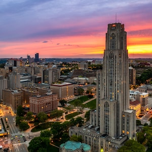 May include: An aerial view of a city skyline at sunset, with a tall, gray stone building in the center. The building has many windows and a pointed roof. The sky is a mix of pink, orange, and purple.