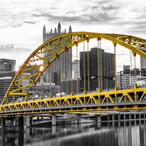 May include: A yellow steel arch bridge spans a river in front of a city skyline. The bridge is in focus, while the city is in black and white.